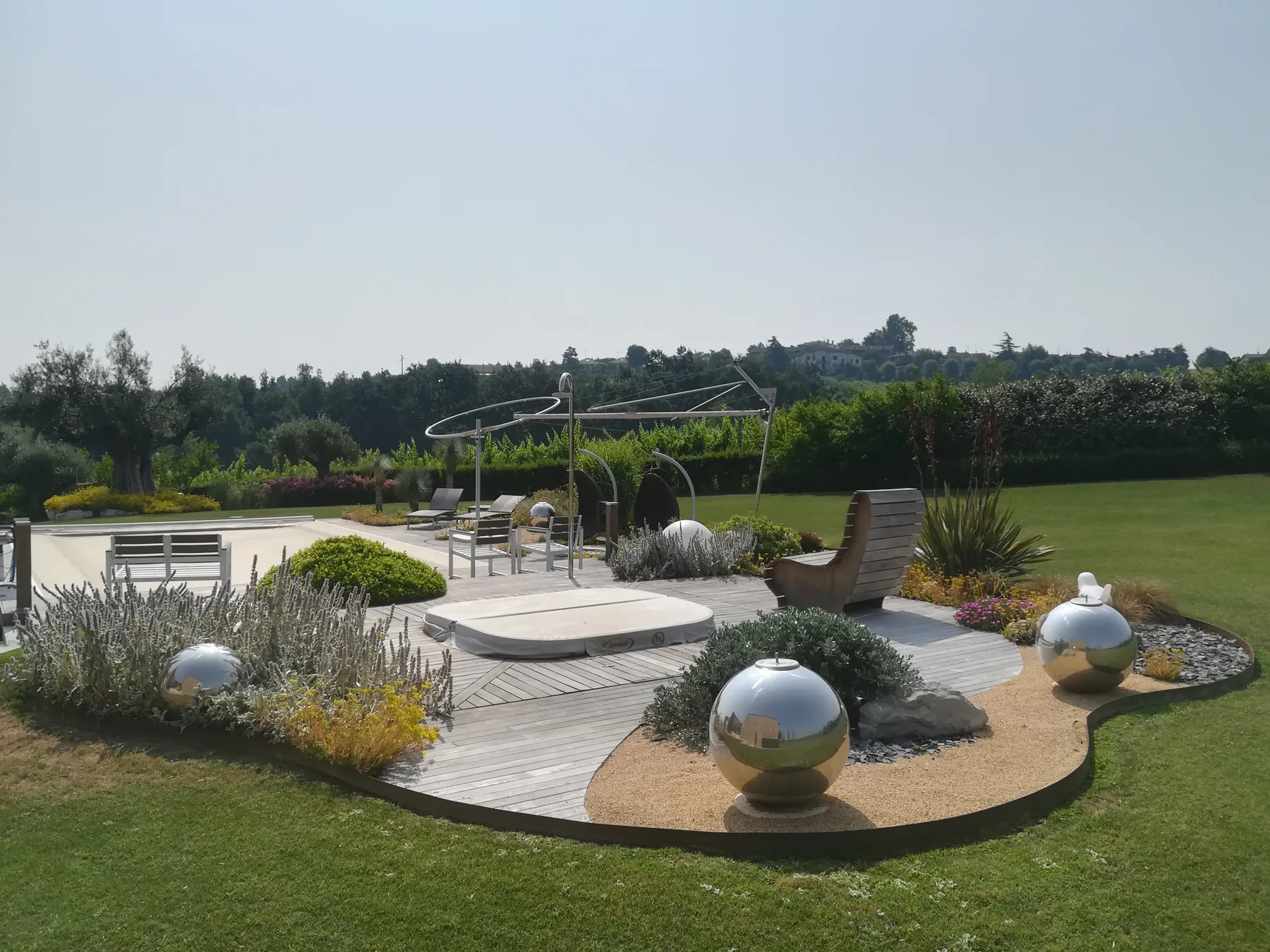 Giardino con pedana in legno grigio a forma libera, sfere in acciaio, cuscini di piante argentate e un prato che arriva fino alla piscina