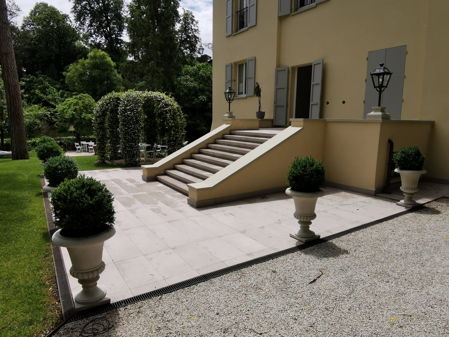 Scalinata d'ingresso di una villa con pavimentazione in pietra chiara, vasi con sfere di bosso, ghiaia e un arco di rampicanti