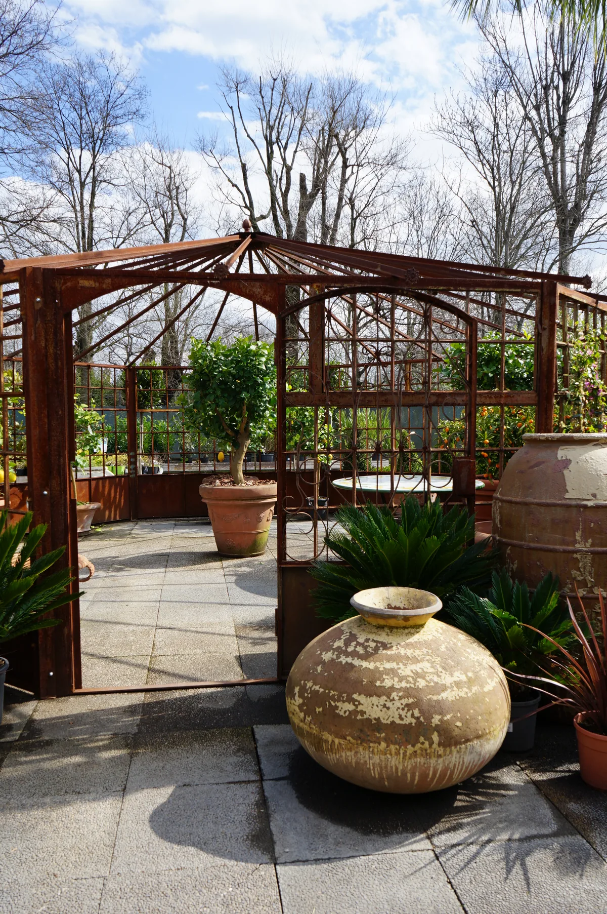 Un gazebo in ferro all'aperto, con un grande orcio di terracotta e piante in vaso sul pavimento di pietra