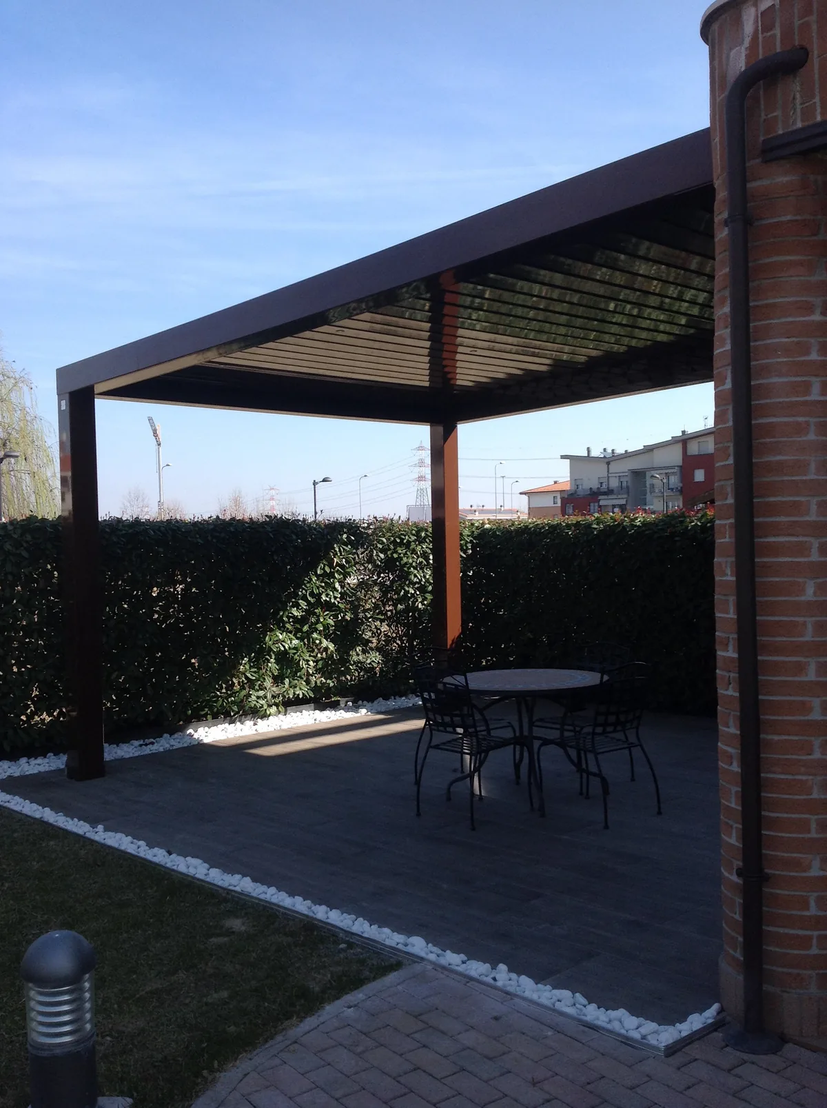 Pergola a lamelle color bruno appoggiata a una colonna in mattoni, con un tavolino rotondo e sedie sotto, davanti a una siepe