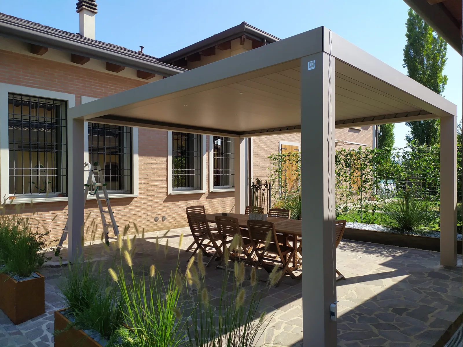 Pergola color tortora davanti a una casa in mattoni, con tavolo e sedie in legno e fioriere in corten piene di graminacee