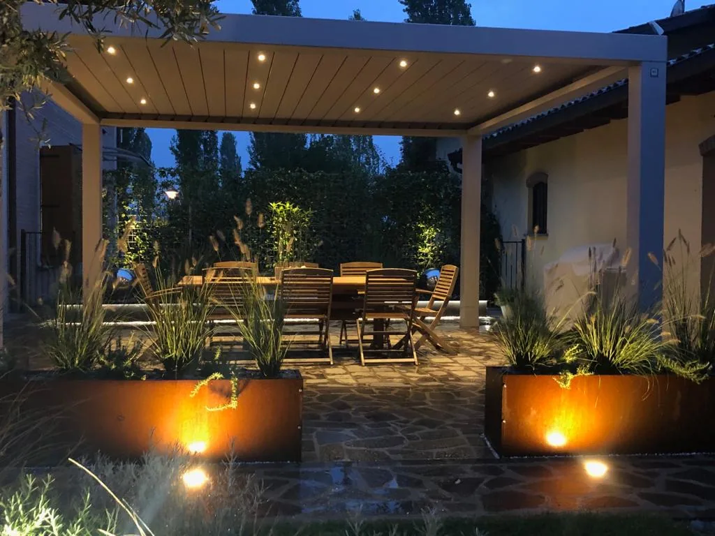 Pergola bioclimatica di sera, con i faretti accesi nel soffitto a lamelle e le fioriere in corten illuminate dal basso