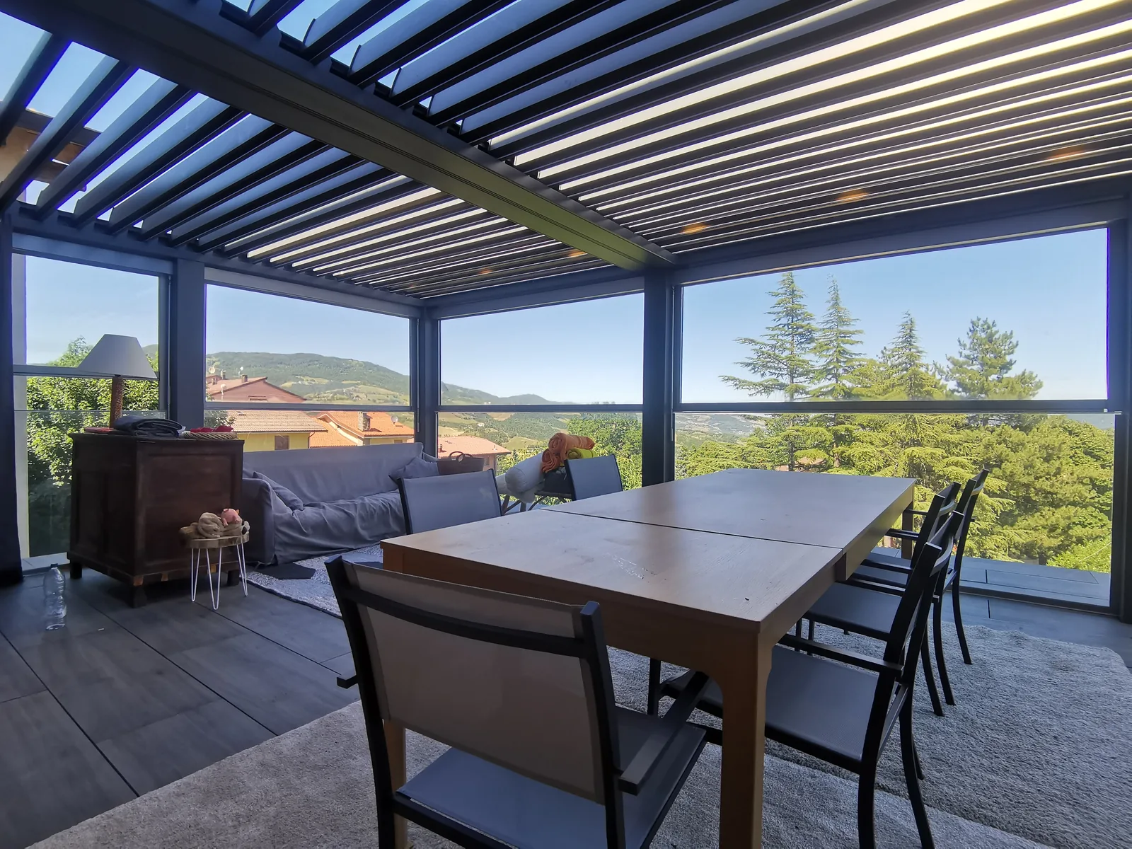 Pergola a lamelle chiusa sui lati da vetrate, con un tavolo e sedie sotto e le colline sullo sfondo