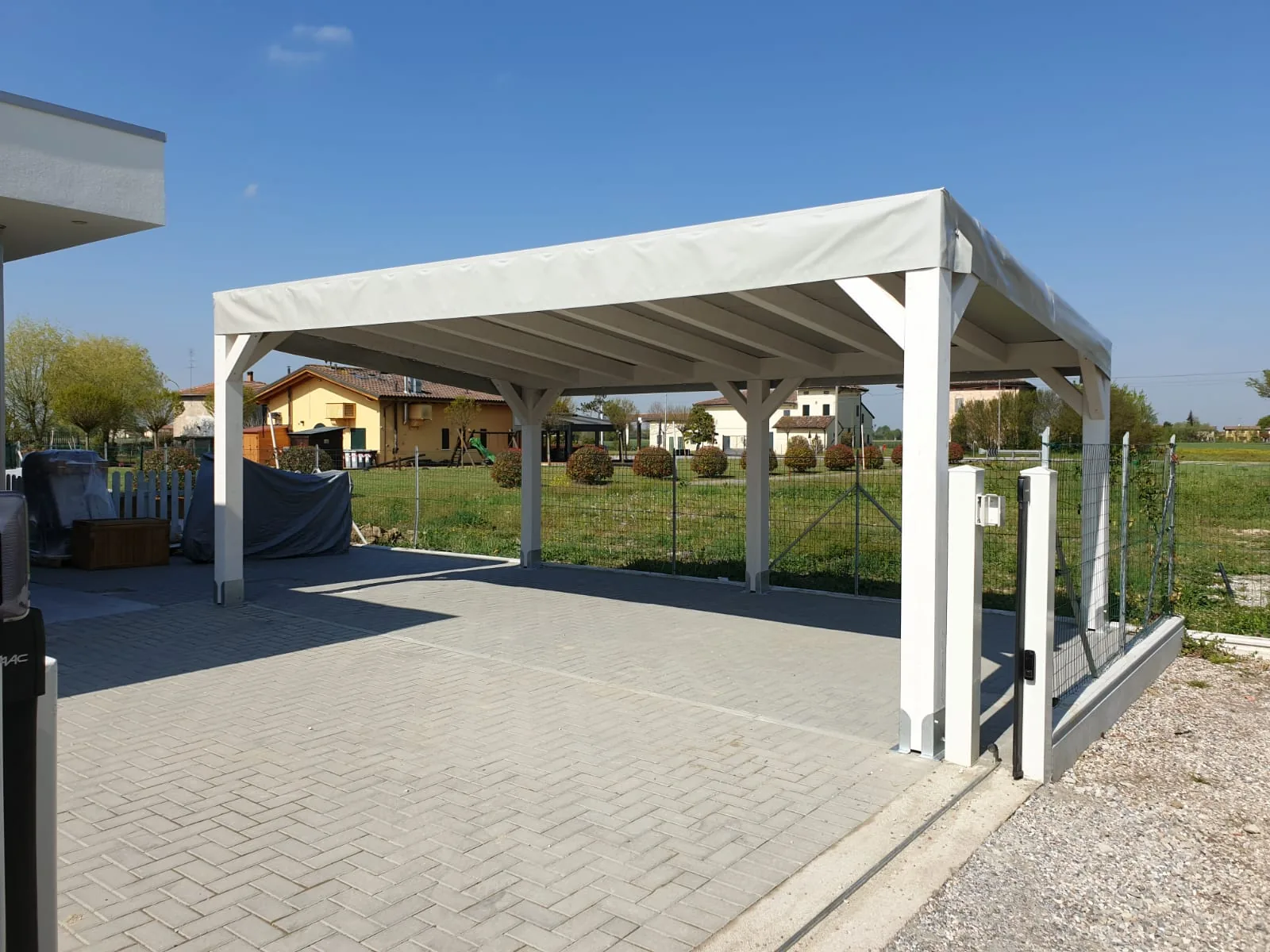 Pergola bianca con copertura in telo chiaro su un cortile in autobloccanti, davanti a un campo