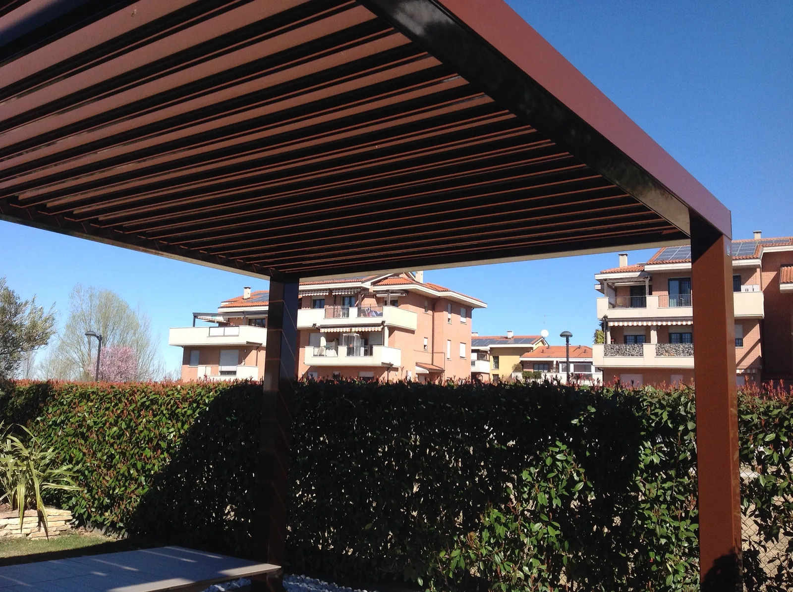 Pergola a lamelle color bruno vista da sotto, con la siepe e le case sullo sfondo