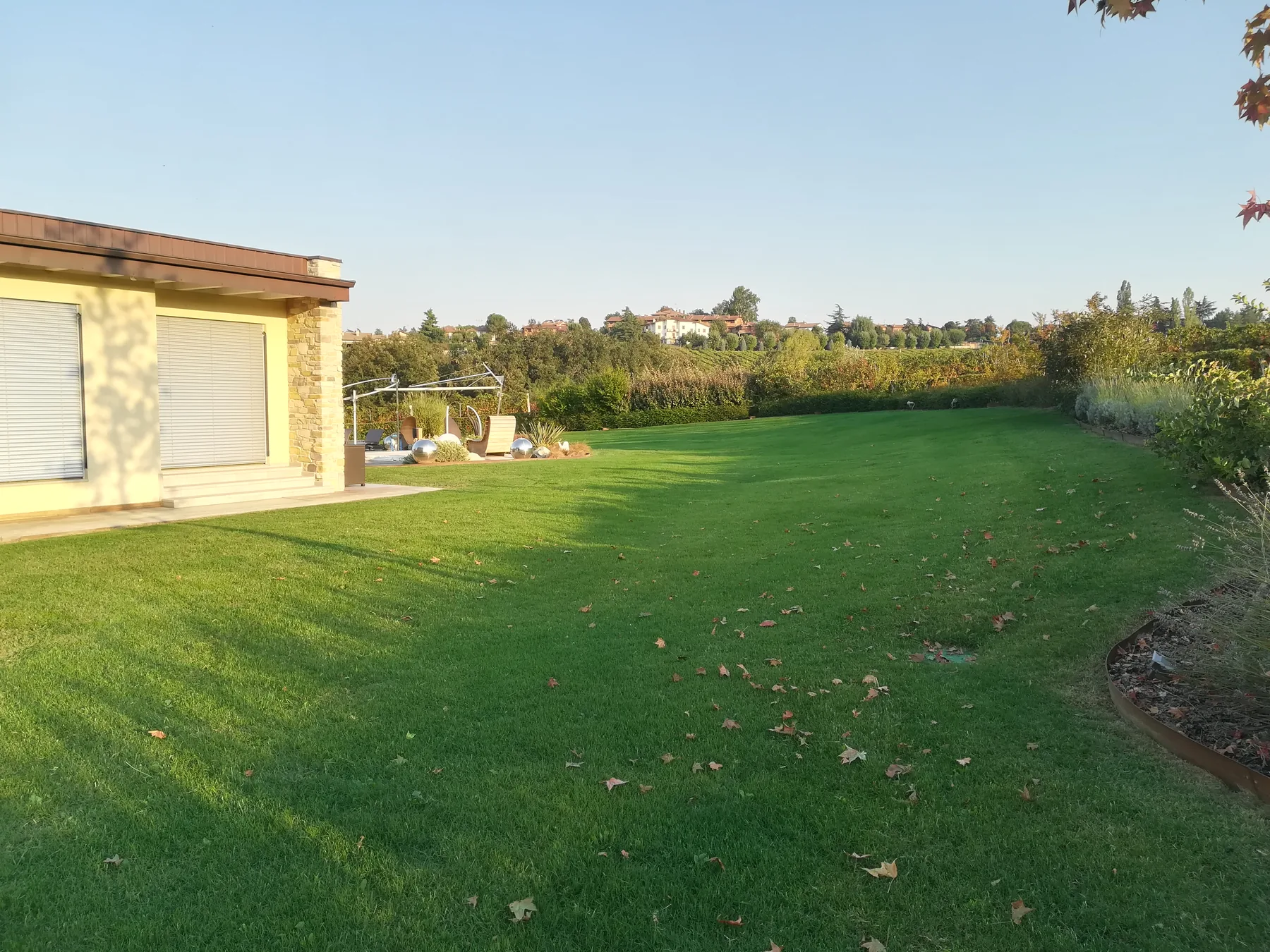 Prato ampio nella luce bassa di fine giornata, con foglie cadute, un'aiuola bordata in acciaio e una casa bassa a sinistra