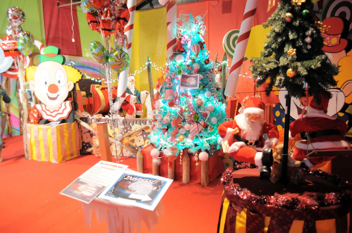 Babbo Natale seduto fra un pagliaccio di cartone, un albero azzurro e i bastoncini di zucchero giganti