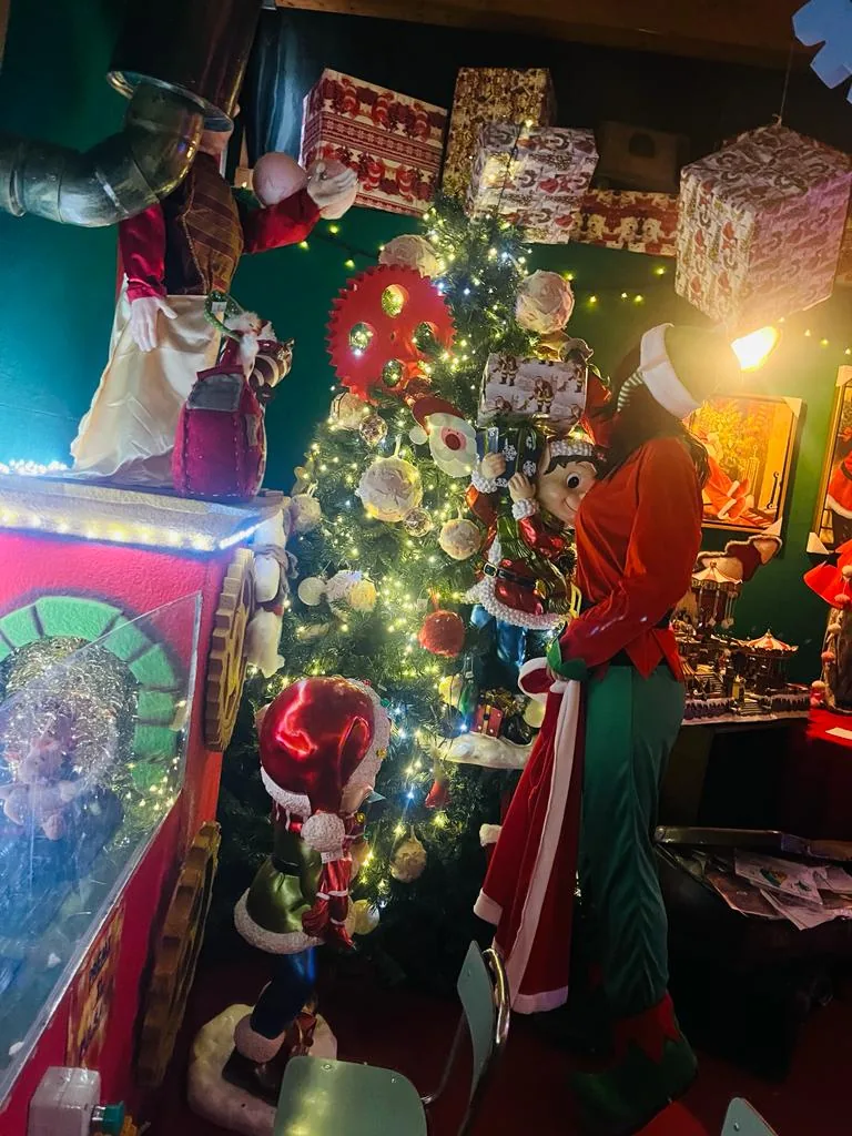 Un elfo accanto all'albero addobbato nel laboratorio di Babbo Natale, sotto i pacchi regalo appesi