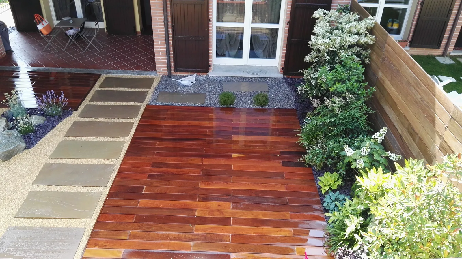 Pedana in listoni di legno scuro appena bagnata, con un camminamento di lastre grigie a lato e aiuole di arbusti lungo una recinzione in legno