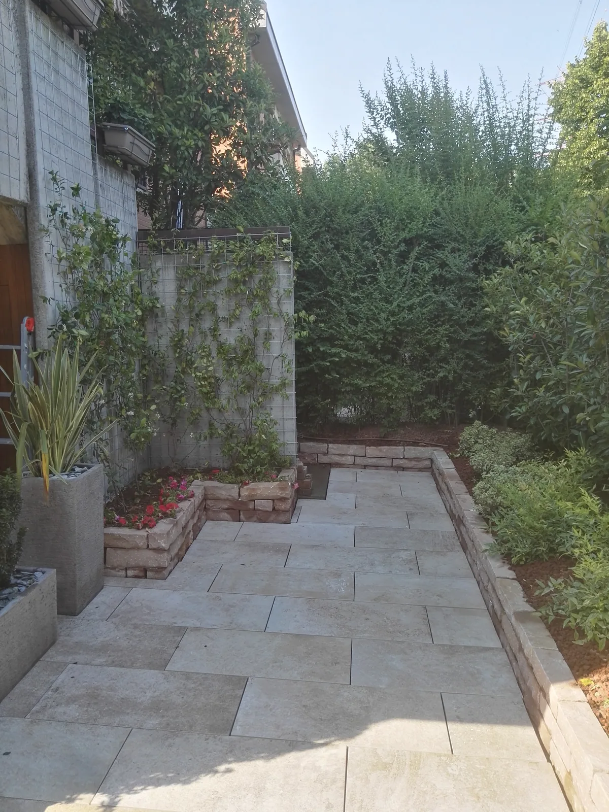 Vialetto in lastre di pietra chiara fra due bordure in mattoni, con piante in vaso e una siepe alta
