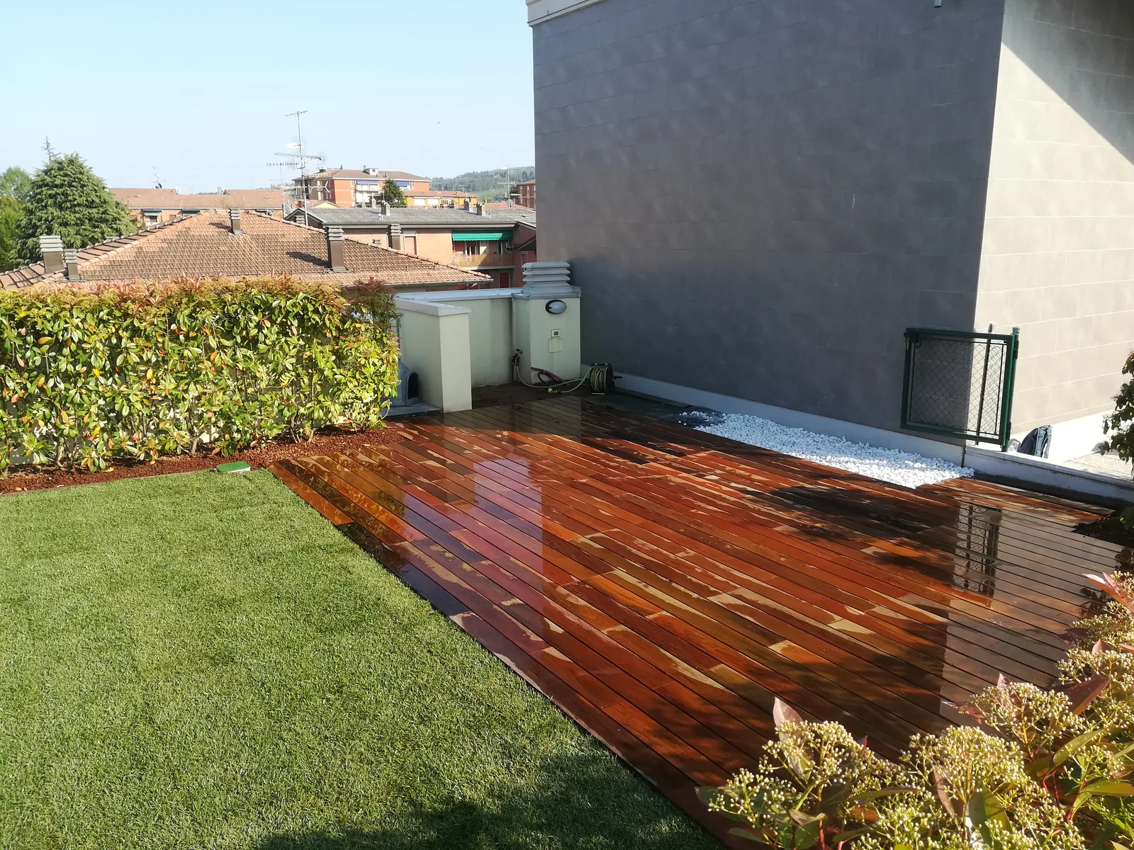 Pedana in legno scuro affiancata a un prato appena posato, su un terrazzo con una siepe di lauro sul bordo