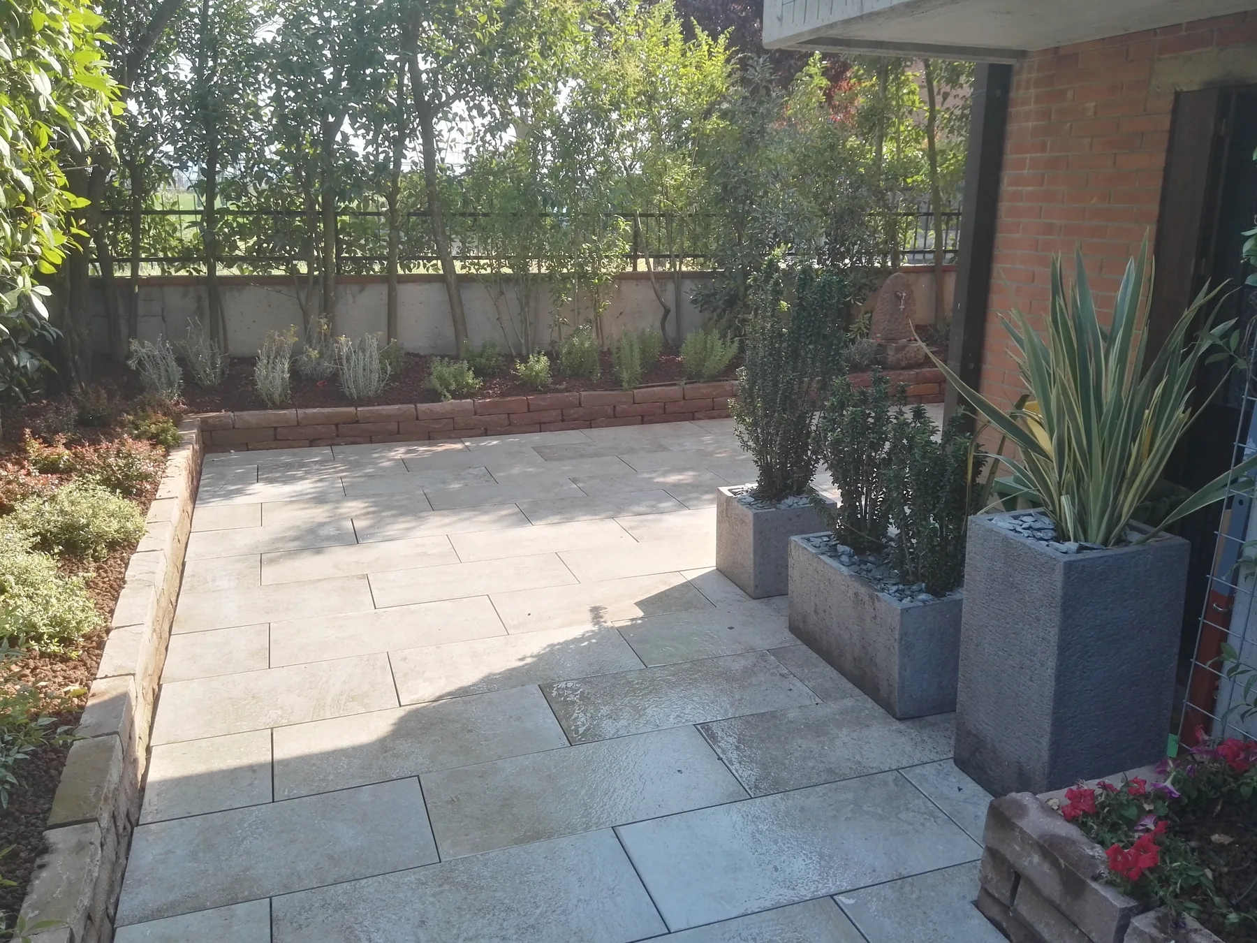 Cortile pavimentato in grandi lastre di pietra chiara, con vasi alti grigi di agavi e phormium e un'aiuola bordata in legno lungo il muro