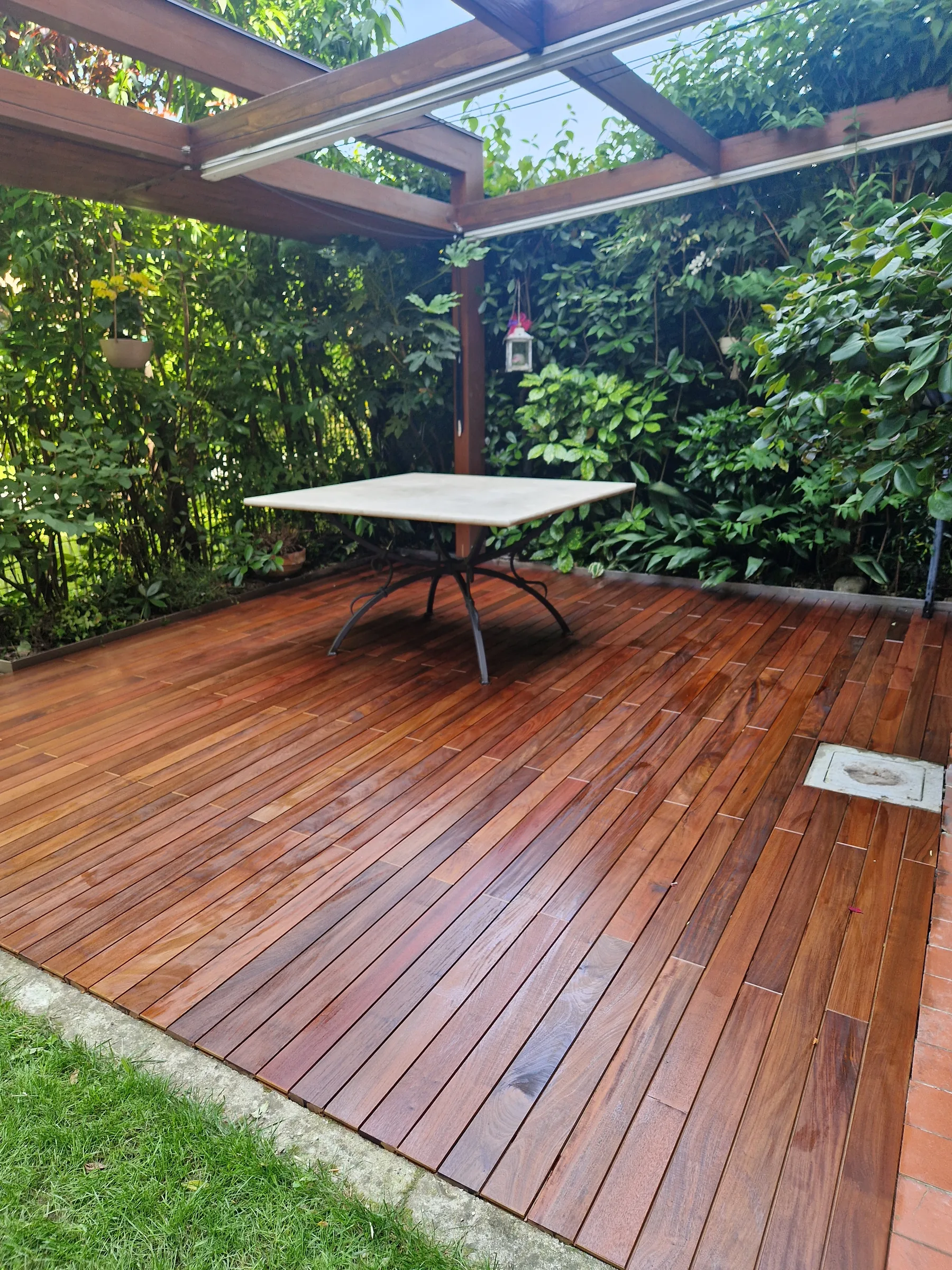 Pedana in listoni di legno sotto una pergola coperta di rampicanti, con un tavolo bianco e il prato sul bordo