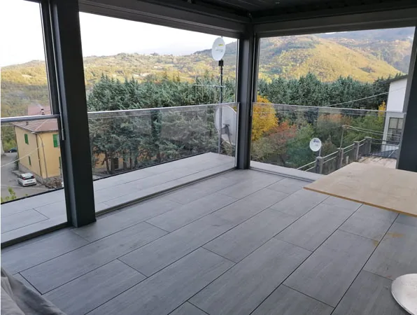 Dopo: dentro la pergola, pavimento porcellanato grigio, pareti vetrate e le colline boscose oltre il parapetto