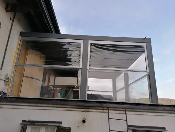 La pergola vista da fuori: struttura grigia con pareti vetrate e tende avvolte, sopra il tetto della casa