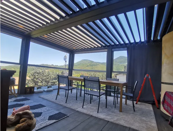 L'interno della pergola: lamelle aperte, pareti vetrate sulle colline, tavolo e sedie sul pavimento porcellanato