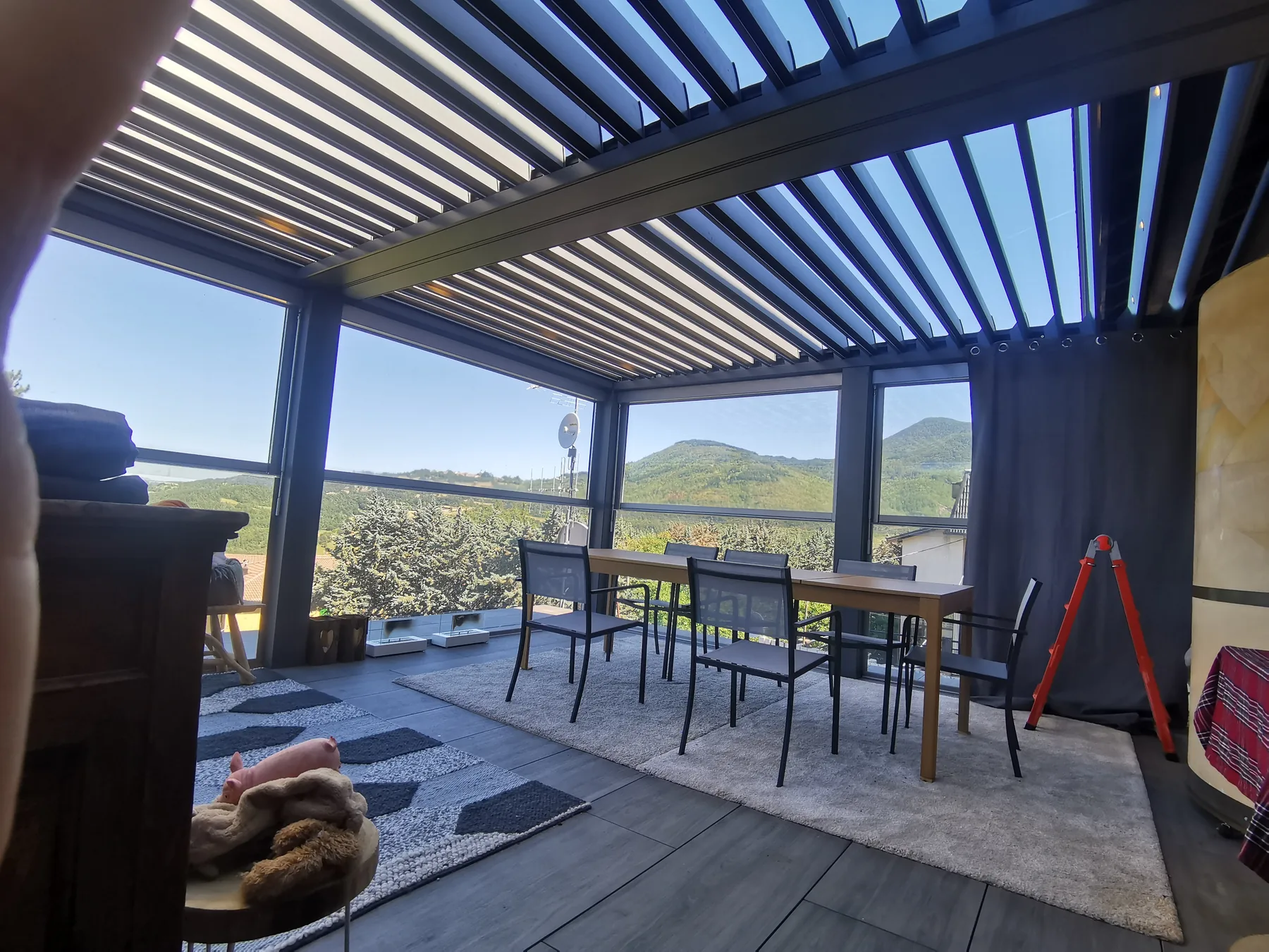 Pergola con le lamelle aperte e le tende scure raccolte, tavolo e sedie sul pavimento porcellanato e le colline oltre le vetrate