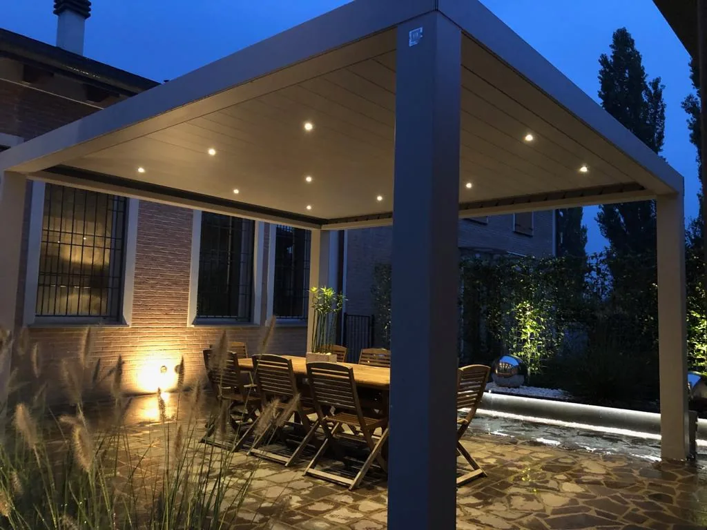 Pergola bioclimatica di sera vista di lato, con i faretti nel soffitto e le graminacee illuminate davanti