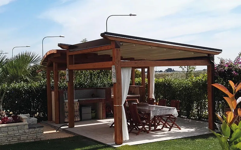 Gazebo in legno con tende bianche raccolte e un tavolo apparecchiato, in mezzo al prato
