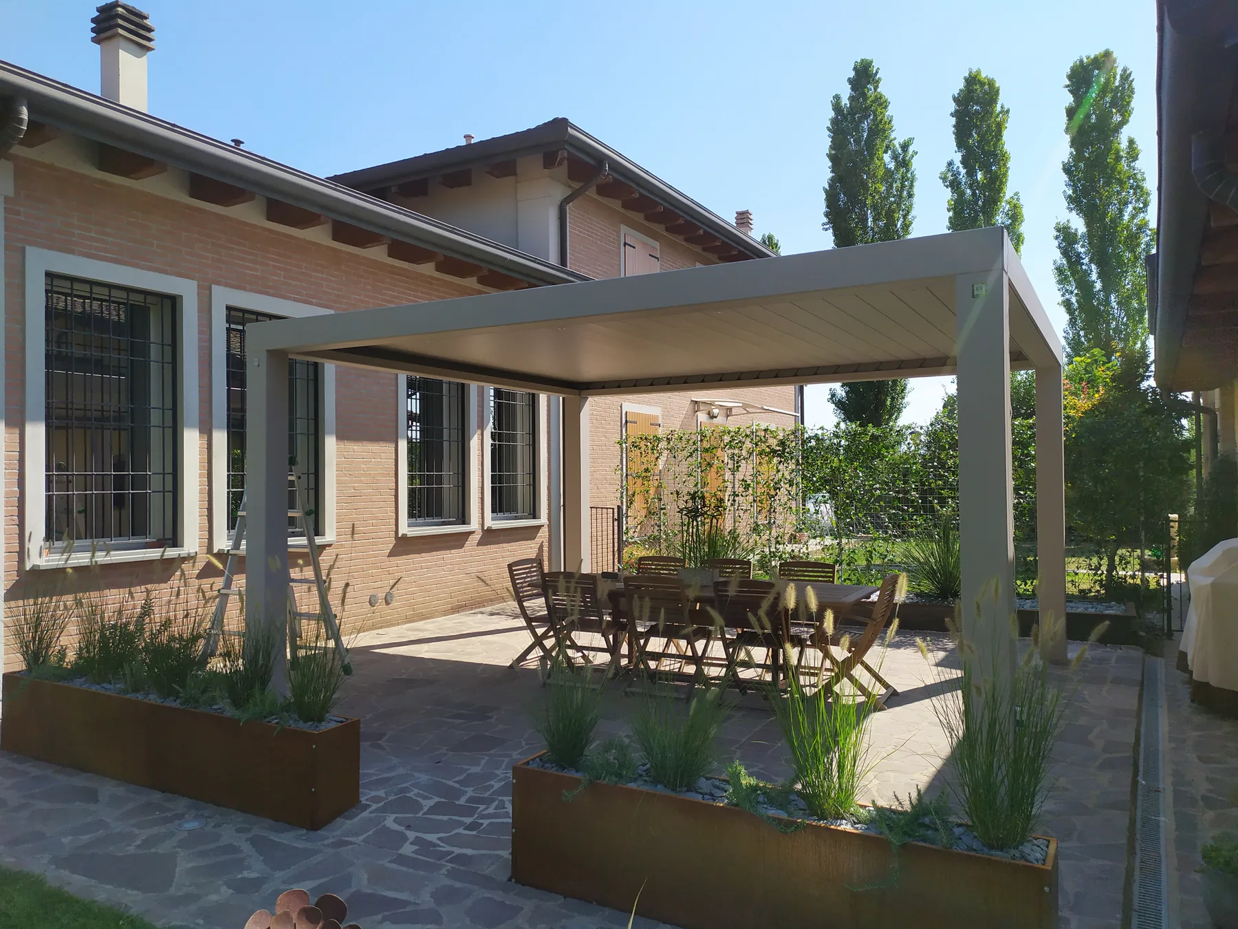 Pergola grigia su un cortile in pietra davanti a una casa di mattoni, con fioriere in corten di graminacee e un tavolo lungo