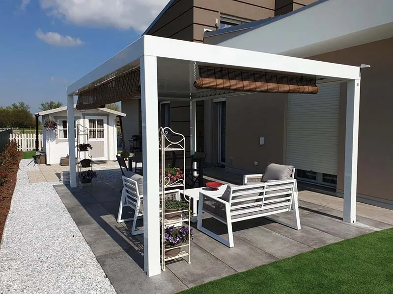Pergola bianca con una tenda avvolgibile sopra un salotto da esterno, su un pavimento in lastre accanto a una casetta in legno