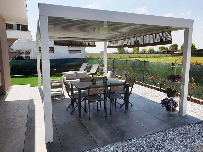 Pergola bianca con tenda avvolgibile sopra un tavolo e un salotto da esterno, al bordo di un prato