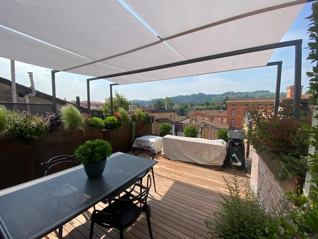 Terrazza in legno sotto una tenda tesa su struttura in ferro, con un tavolo lungo e le fioriere verso i tetti