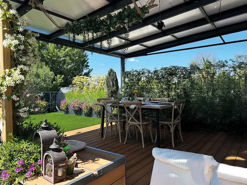 Pergola con copertura trasparente e gelsomino, sopra una pedana in legno con il tavolo apparecchiato e le fioriere