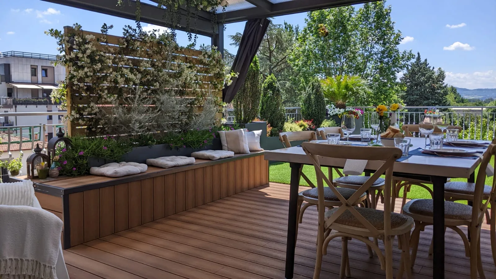 Terrazza in legno con panca imbottita lungo le fioriere, un tavolo apparecchiato e un pannello di rampicanti