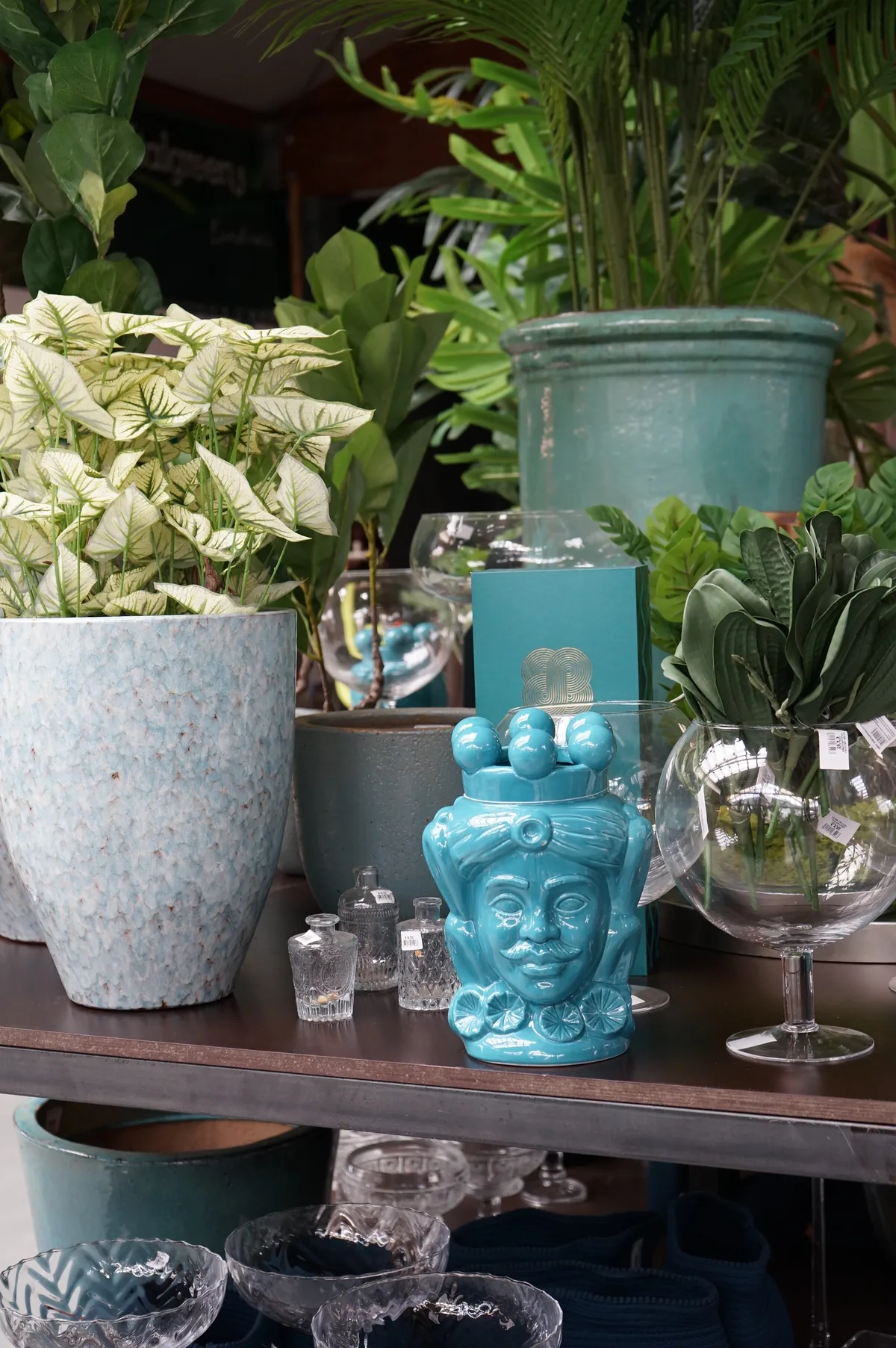 Portavasi in cemento chiaro e in ceramica verde acqua con piante a foglia variegata, un vaso a testa azzurro e bicchieri di vetro