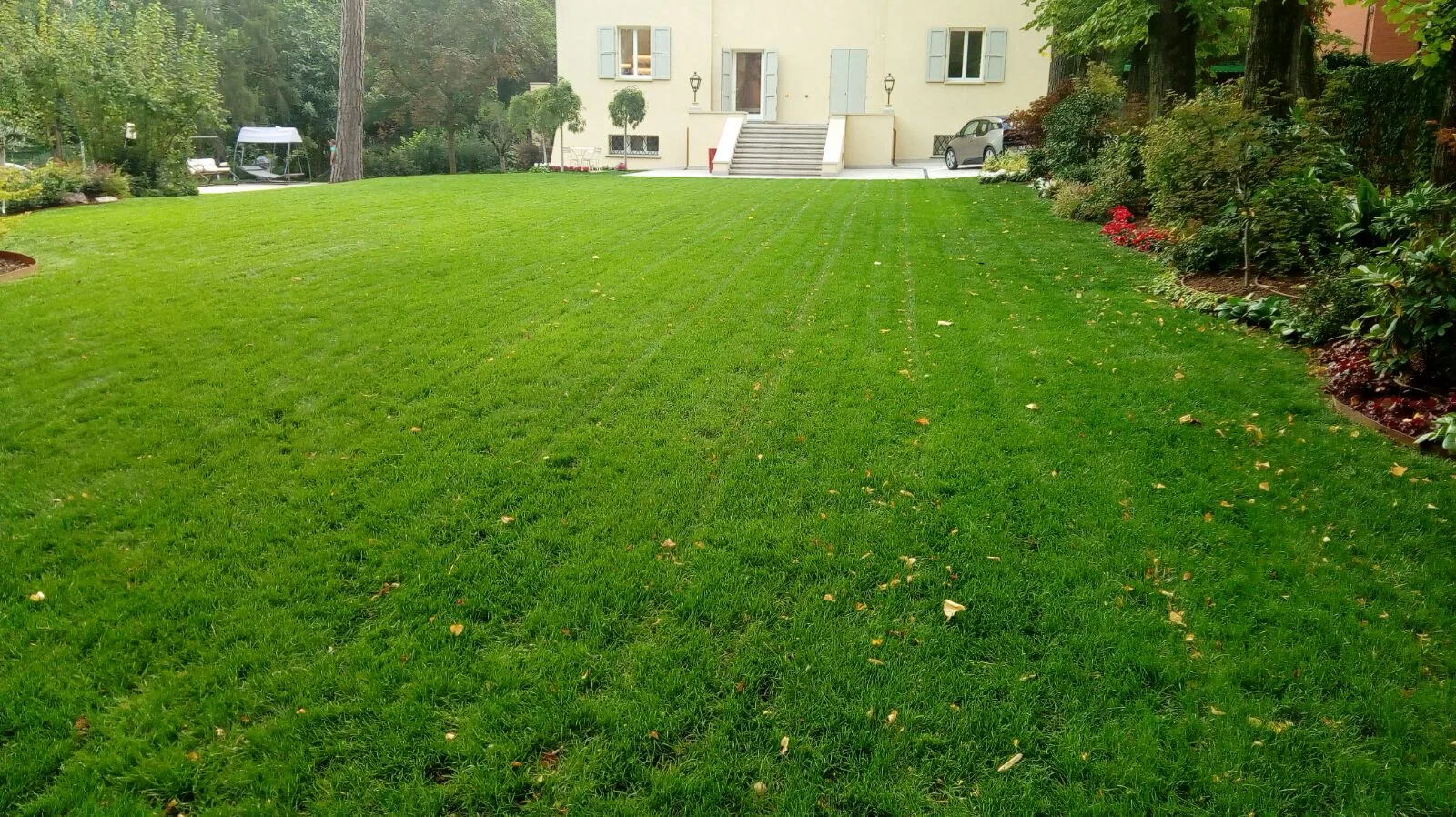 Prato rasato con le strisce del taglio davanti a una villa chiara, con la bordura fiorita sulla destra