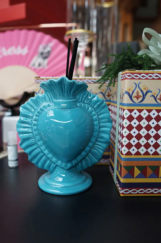 Vaso in ceramica turchese a forma di cuore accanto a una scatola decorata a motivi geometrici e a un ventaglio rosa