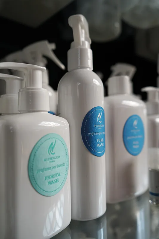Flaconi bianchi con erogatore spray e etichette azzurre, allineati su un ripiano di vetro