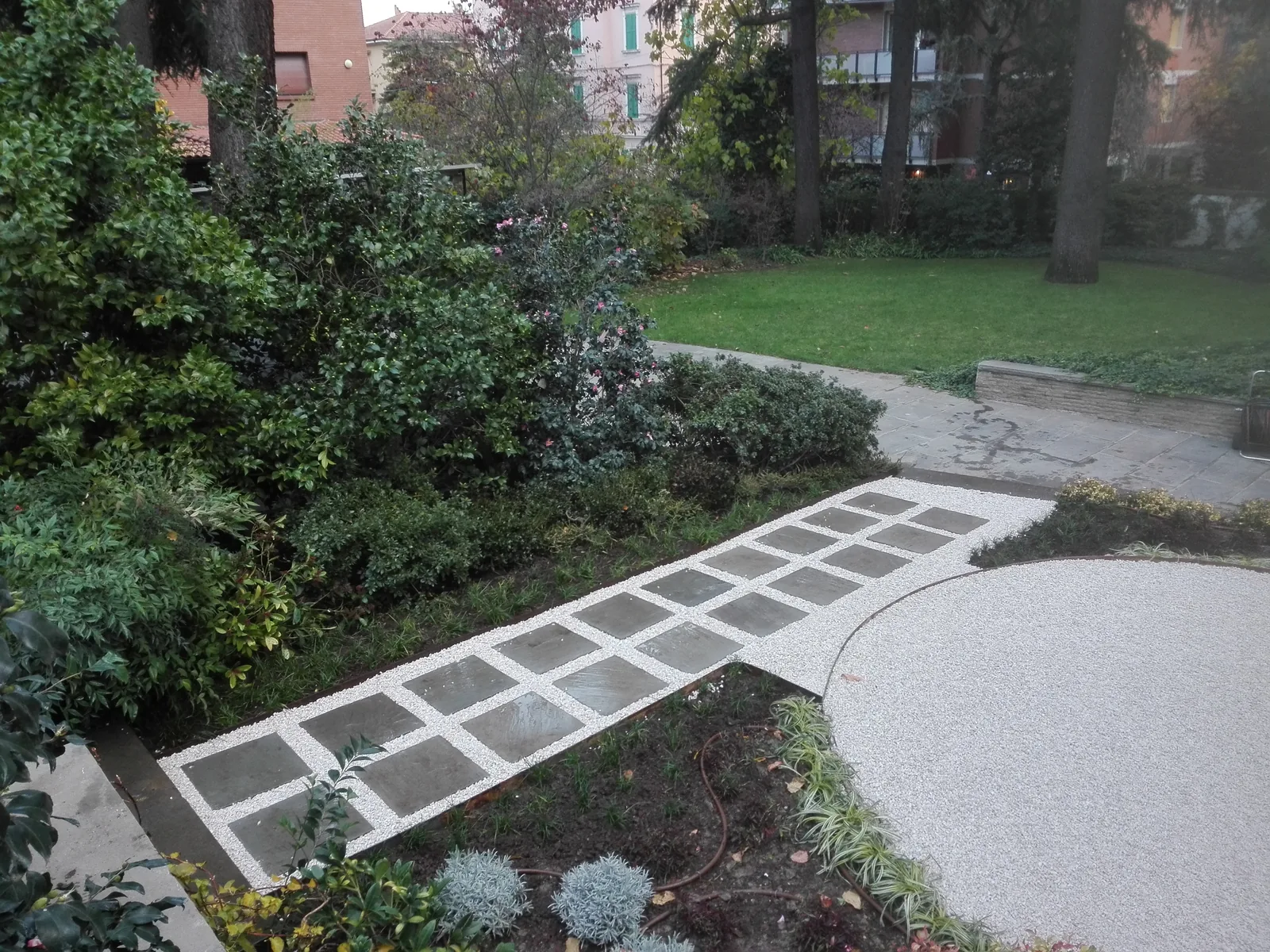 Camminamento in lastre quadrate con fughe di ghiaia fra aiuole di arbusti, che porta a un'area di ghiaietto chiaro