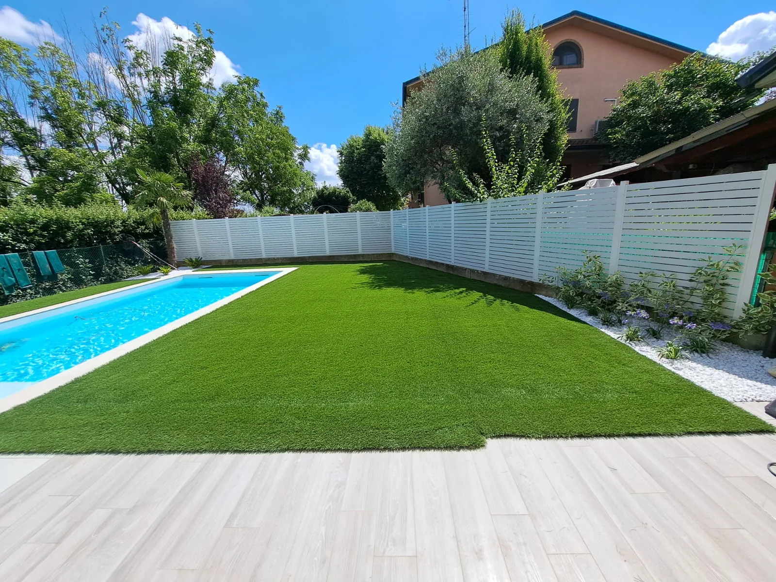 Prato sintetico lungo una piscina, chiuso da una staccionata bianca, con un olivo e una casa sul fondo