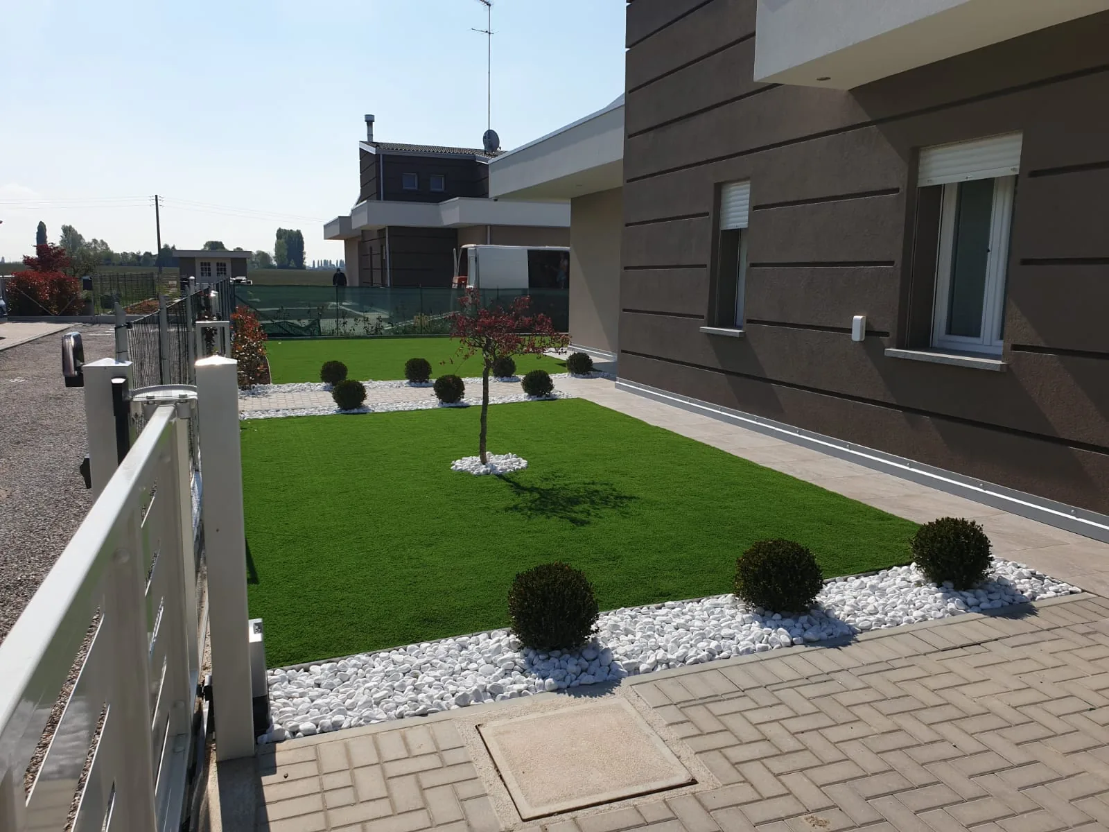 Prato sintetico nel giardino di una casa, con sfere di bosso su ciottoli bianchi, un alberello e una pavimentazione a spina di pesce