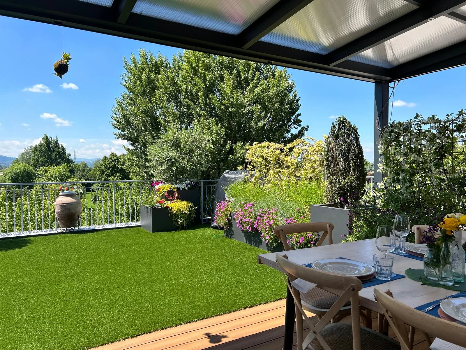 Prato sintetico su una terrazza coperta, con la tavola apparecchiata, fioriere fiorite e il verde oltre la ringhiera