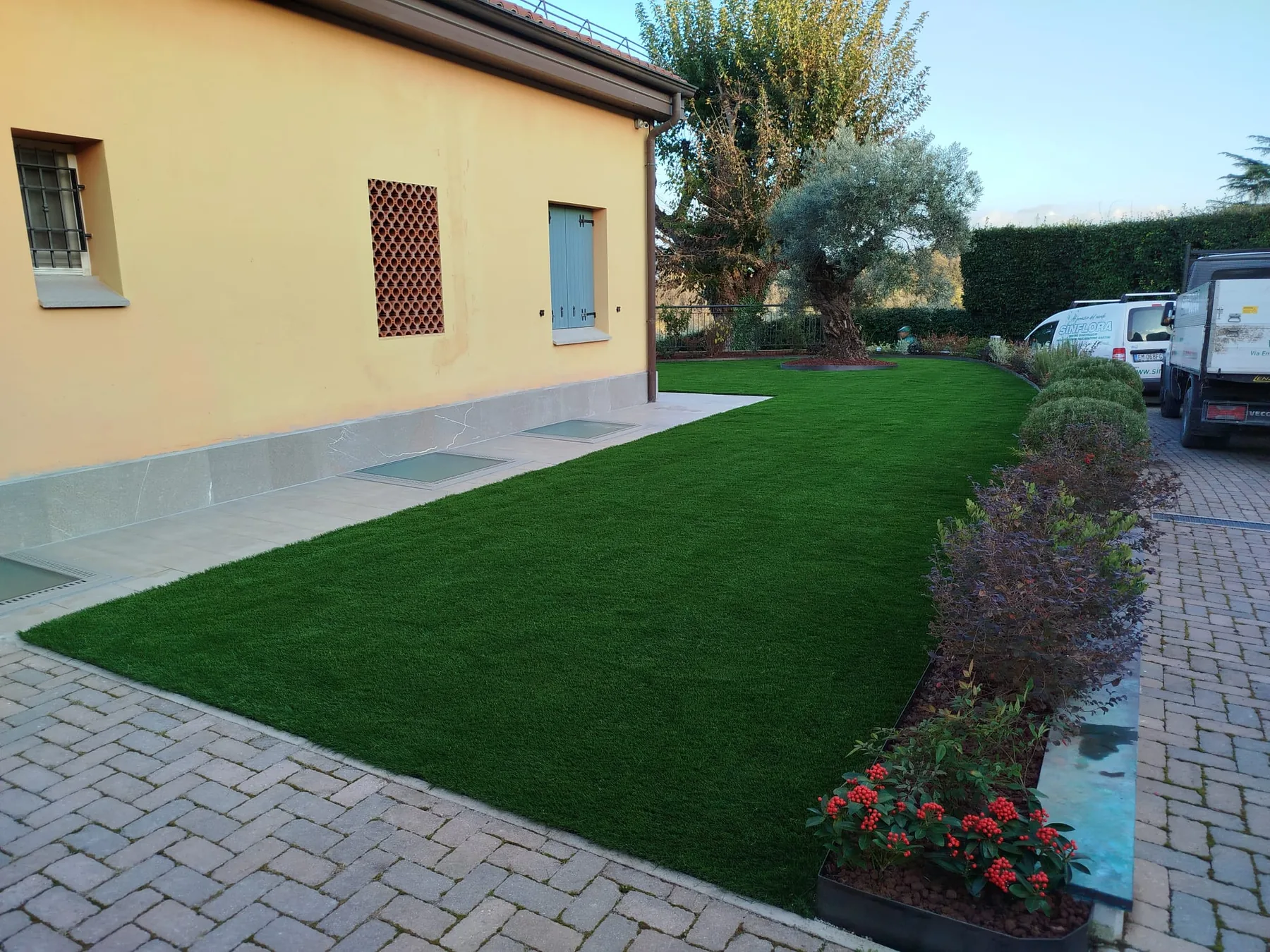 Prato sintetico lungo una casa gialla, bordato da un'aiuola di arbusti e fiori rossi accanto a un vialetto in autobloccanti