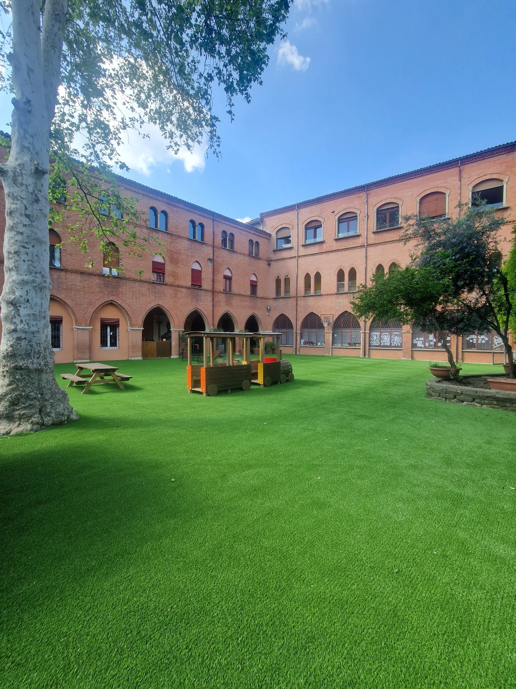 Cortile di un edificio in mattoni con portico ad archi, coperto di prato sintetico, con tavoli da pic-nic e giochi in legno