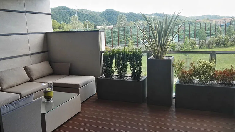 Angolo salotto su un terrazzo in legno, con fioriere scure di cipressi e le colline oltre il parapetto
