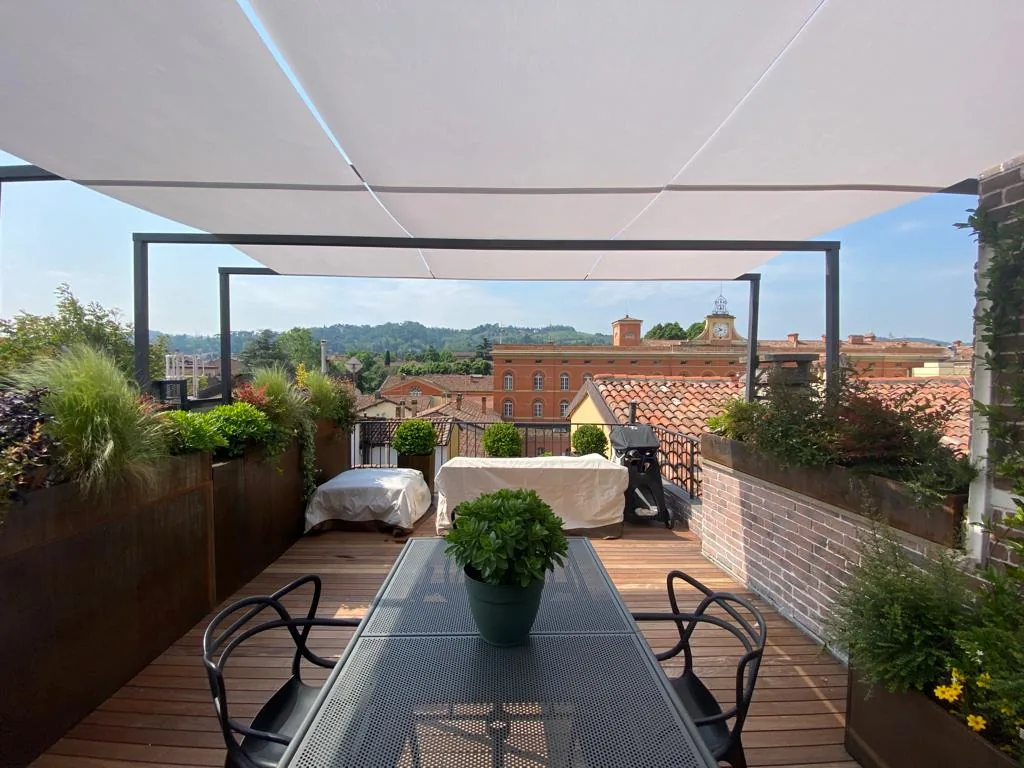 Tavolo in rete metallica sotto la tenda bianca, fra le fioriere in corten e il muro in mattoncini, con i tetti e le colline sul fondo