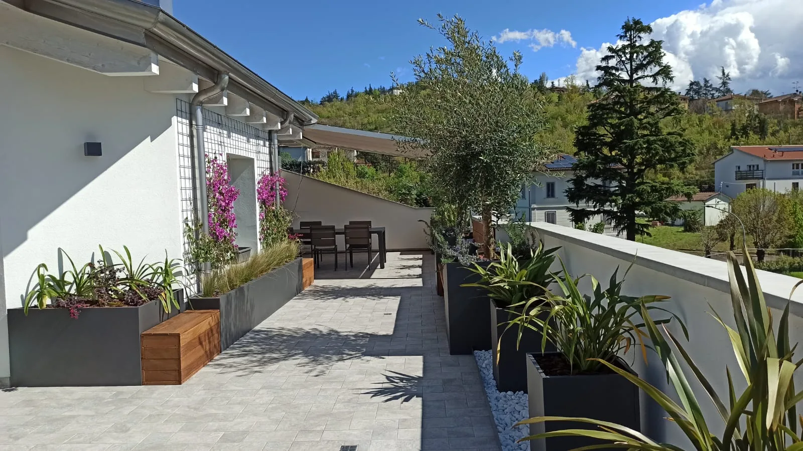 Terrazzo lungo con fioriere antracite, una bouganville in fiore, un olivo e una panca in legno, con le colline alle spalle