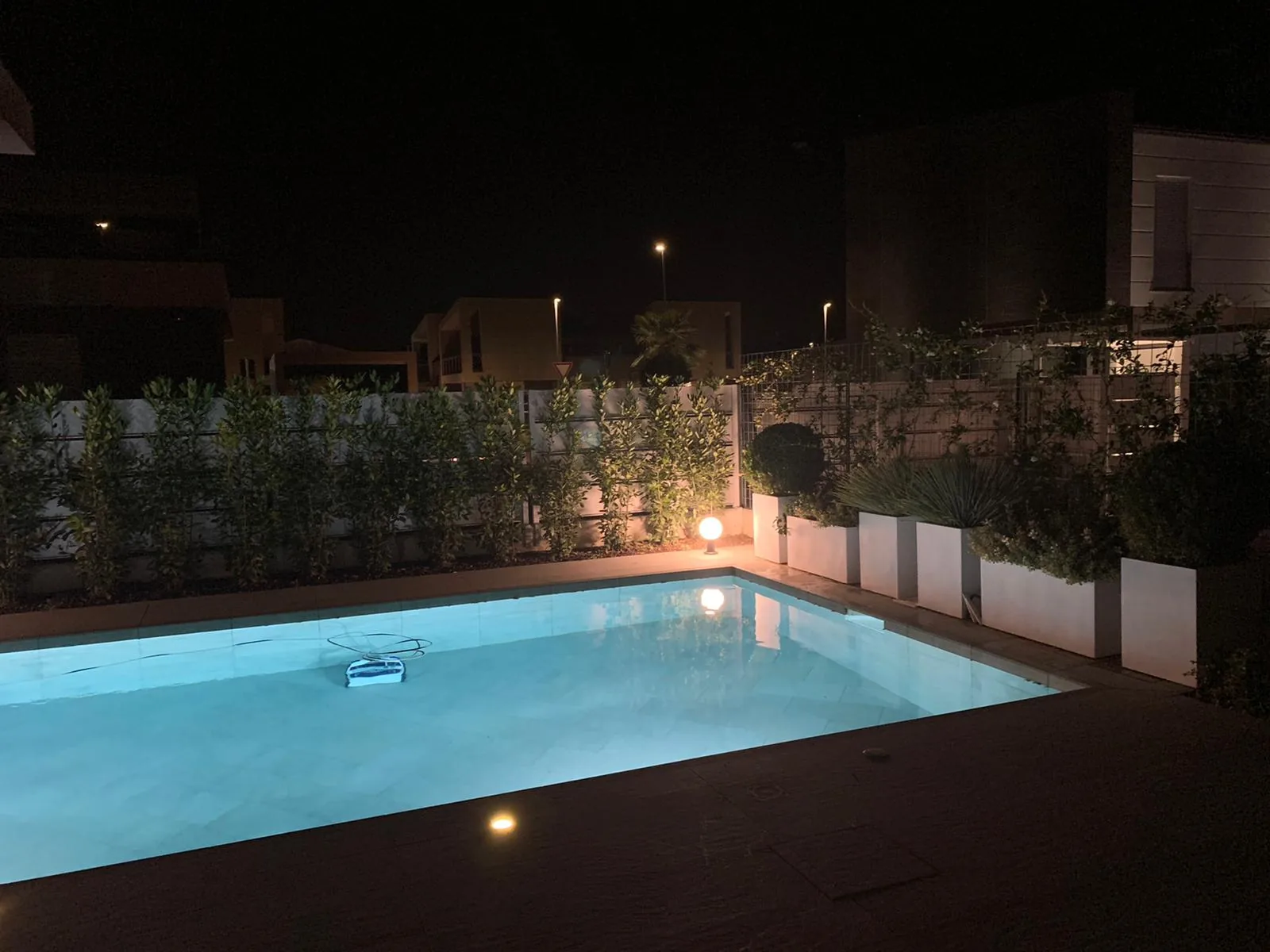 Piscina illuminata di notte, con la siepe e le fioriere bianche lungo il bordo accese da lampade