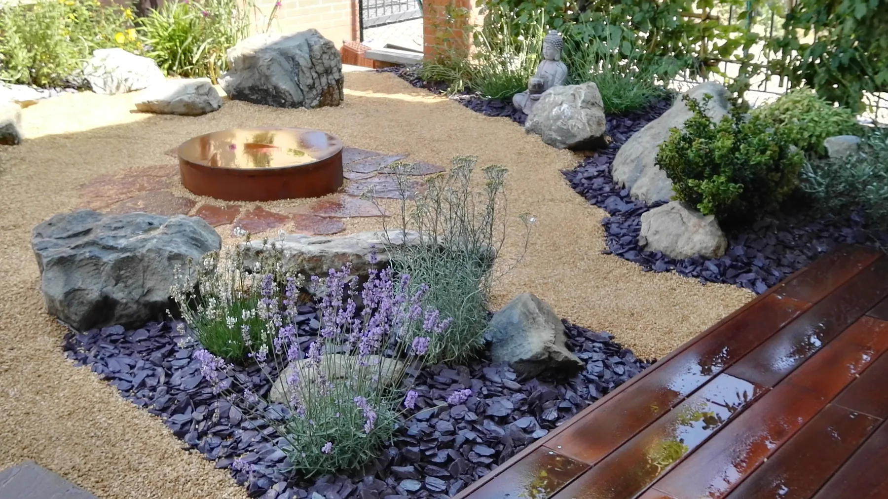 Aiuola secca con ghiaia dorata, scaglie di ardesia viola, massi, lavanda e una vasca rotonda in corten accanto a una pedana in legno