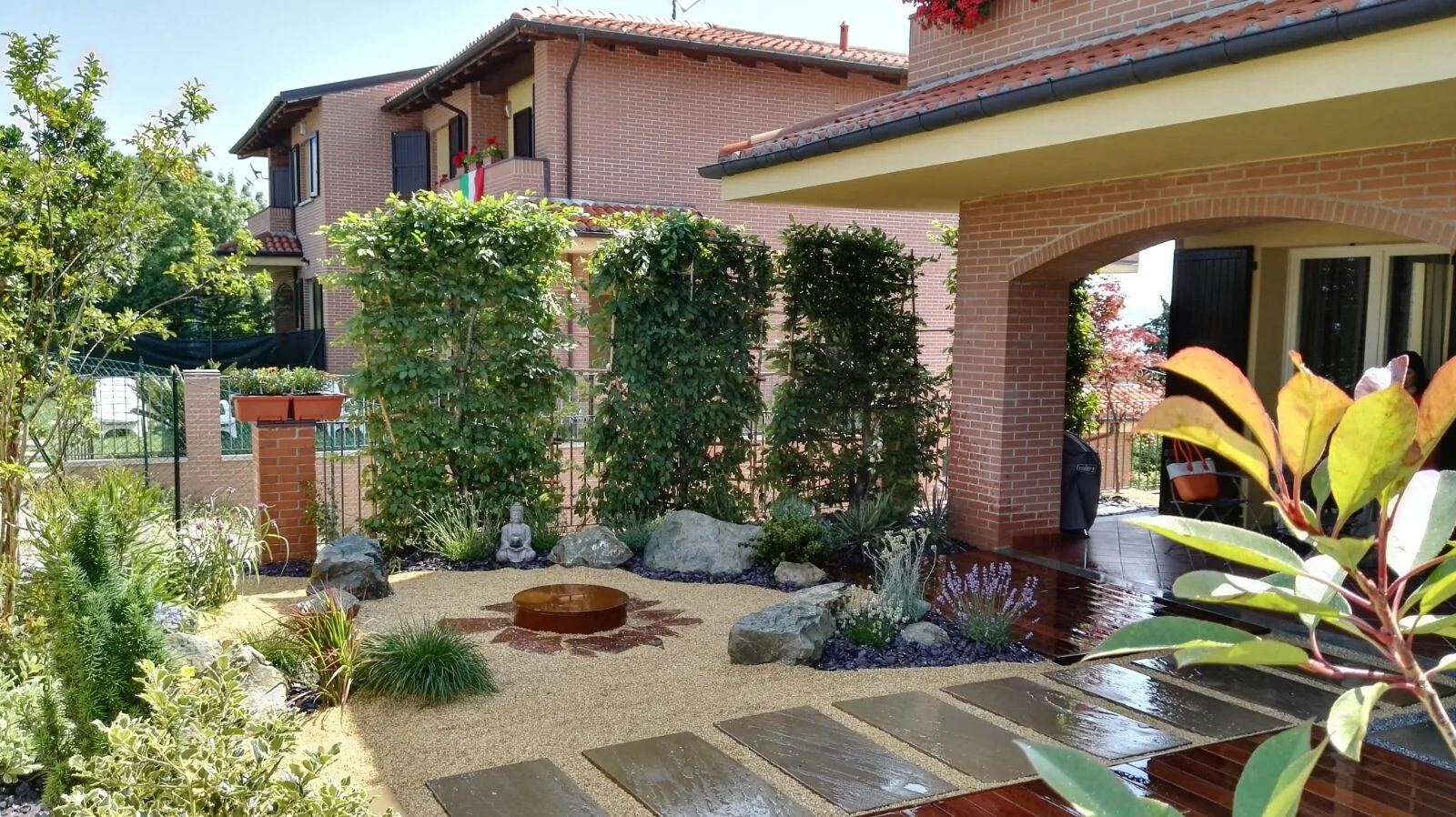 Cortile d'ingresso con passi in pietra sulla ghiaia, massi, una vasca rotonda in corten e grigliati coperti di rampicanti