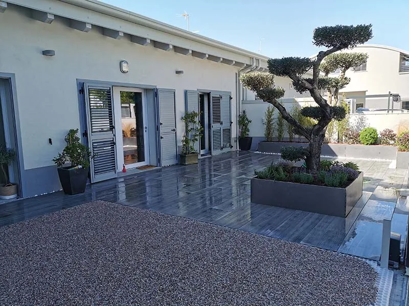 Terrazzo lastricato con un albero potato a nuvola in una grande fioriera grigia e piante in vaso lungo la casa