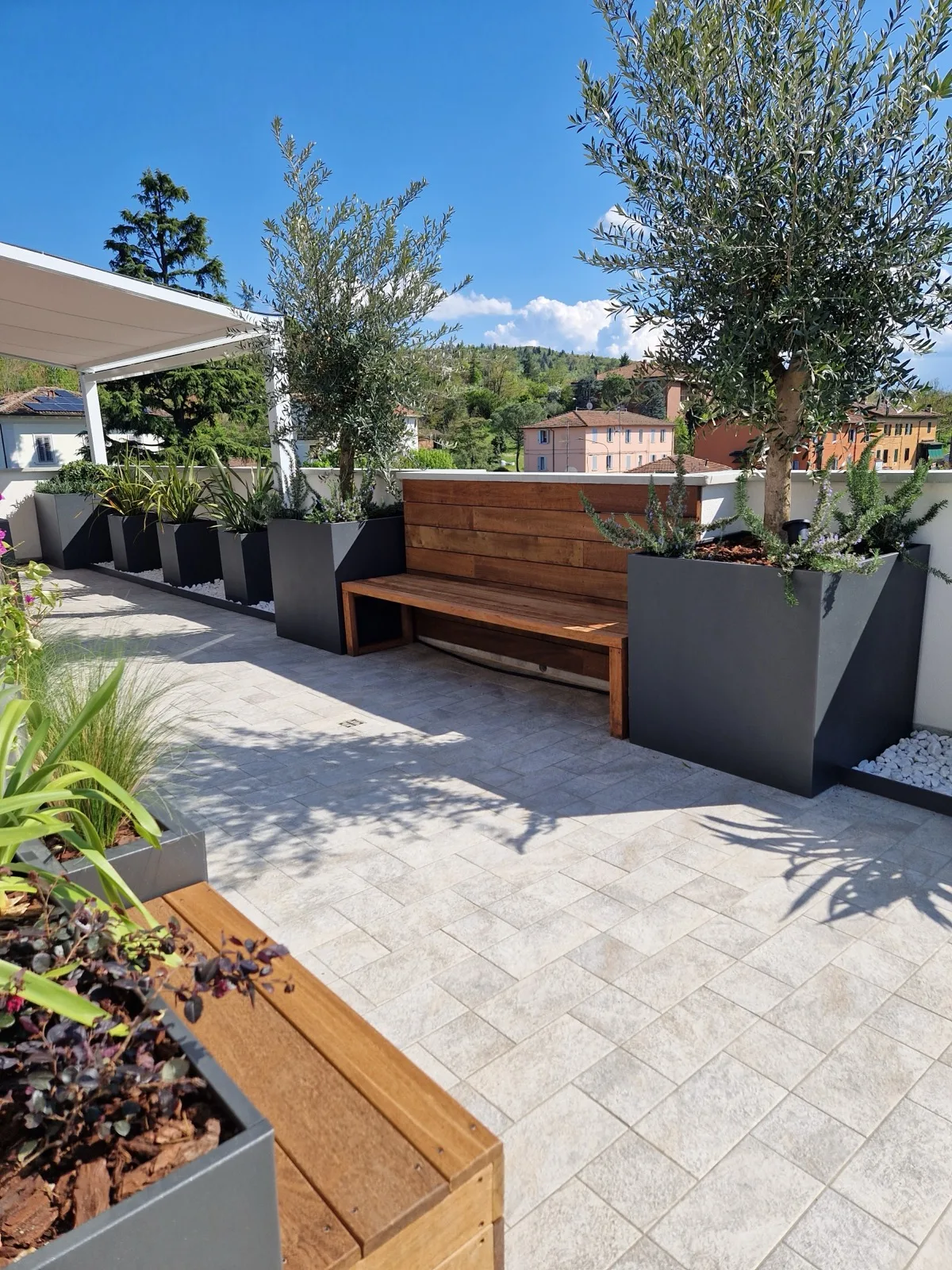 Terrazza con fioriere antracite e in legno, panca a tutta lunghezza, olivi e agavi, con le colline sullo sfondo