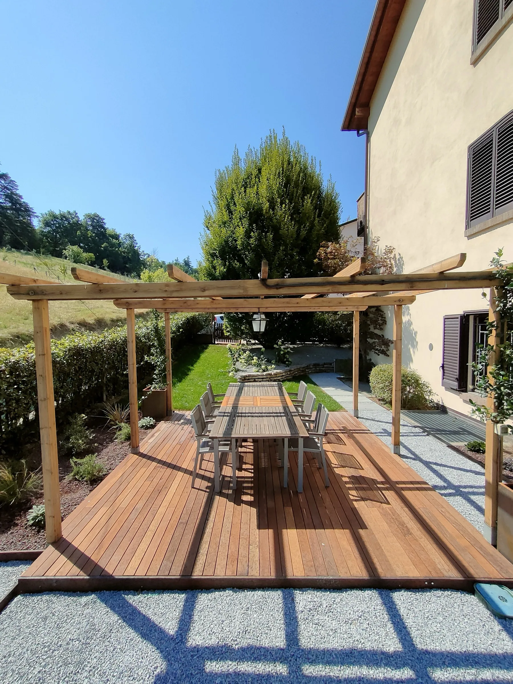 Pergola in legno a travi incrociate sopra un tavolo lungo su pedana in listoni, con vialetto in ghiaia e prato ai lati