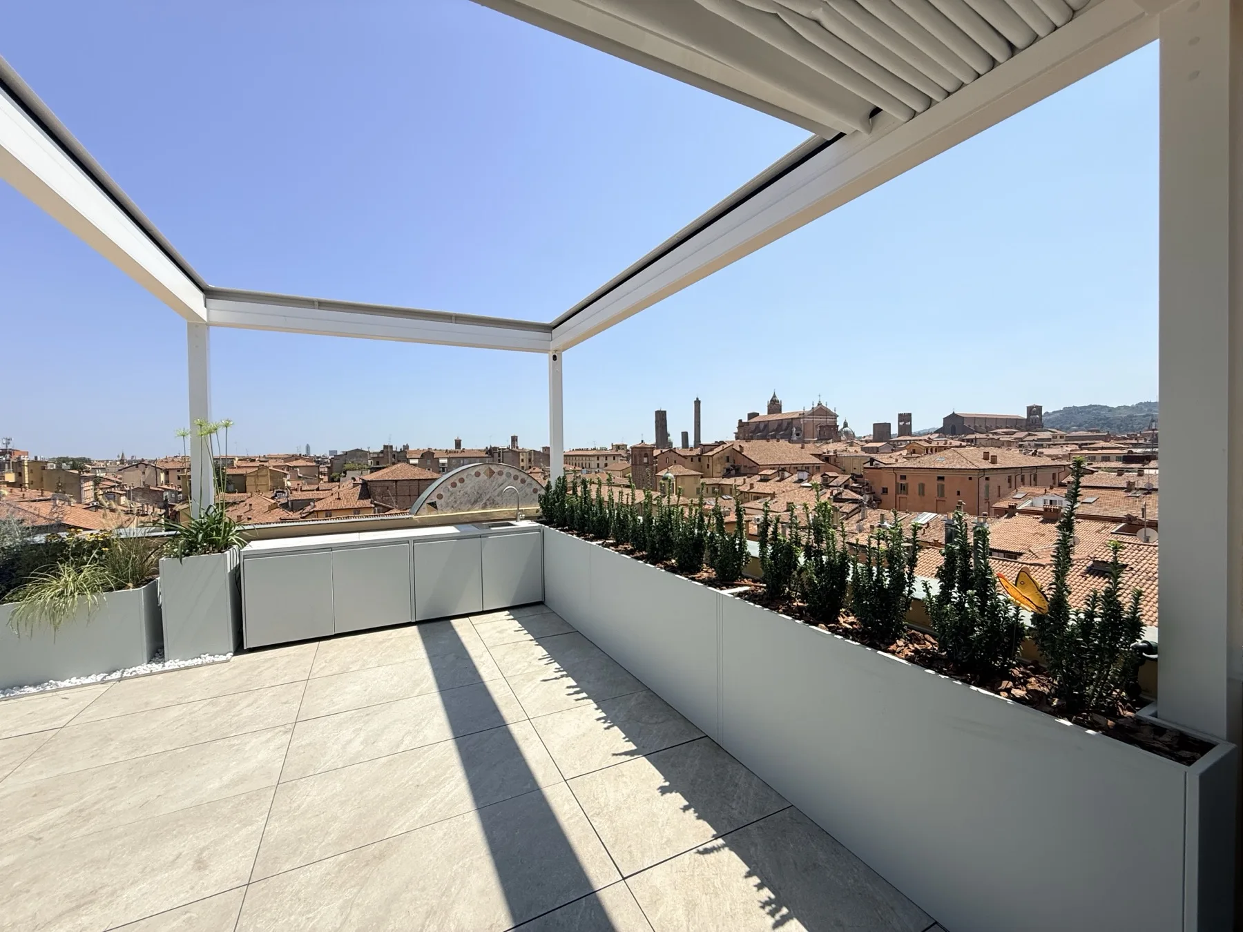 Terrazza all'ultimo piano con pergola bioclimatica a lamelle, fioriere continue di cipressi nani e i tetti della città con le due torri sul fondo