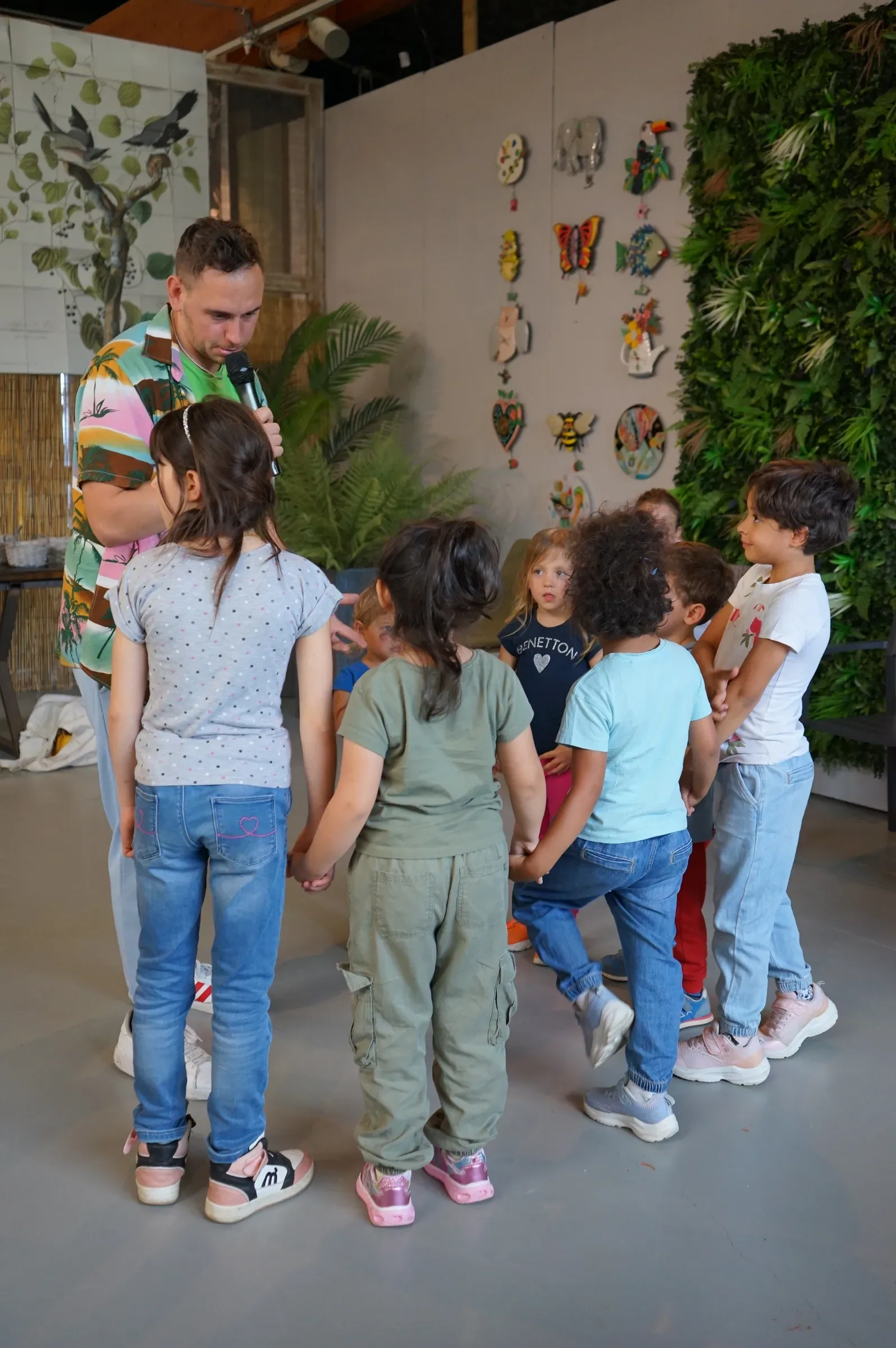 Un animatore con il microfono in piedi accanto ai bambini riuniti in cerchio, davanti alla parete di verde